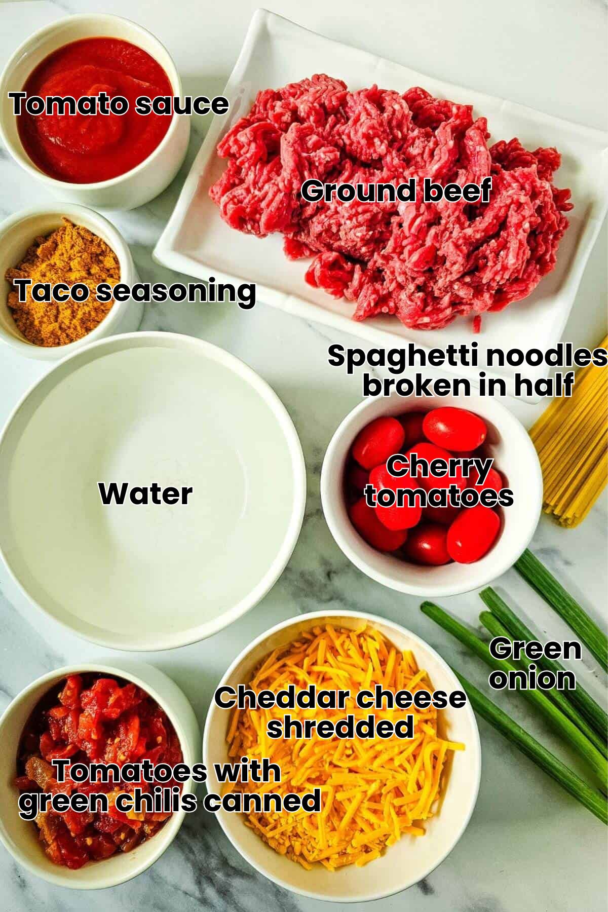 Ingredients on a countertop include ground meat, cherry tomatoes, shredded cheese, diced tomatoes, taco seasoning, tomato sauce, green onions, water, and uncooked spaghetti.