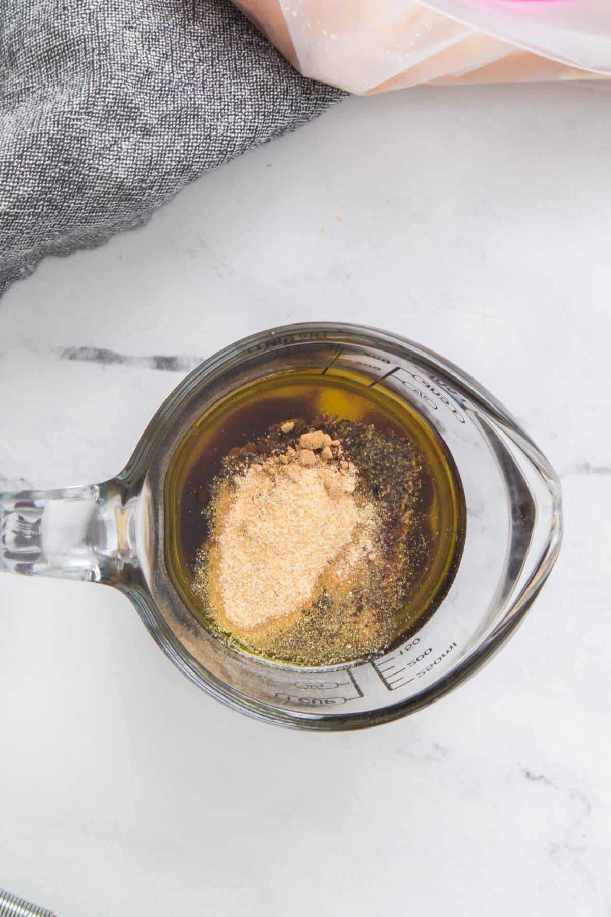 A measuring cup on a table contains olive oil and various powdered spices.