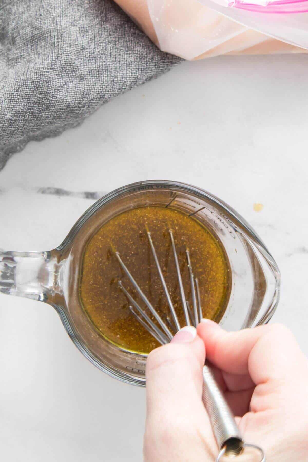 Hand whisking brown marinade in a clear measuring cup on a marble surface next to a gray cloth.