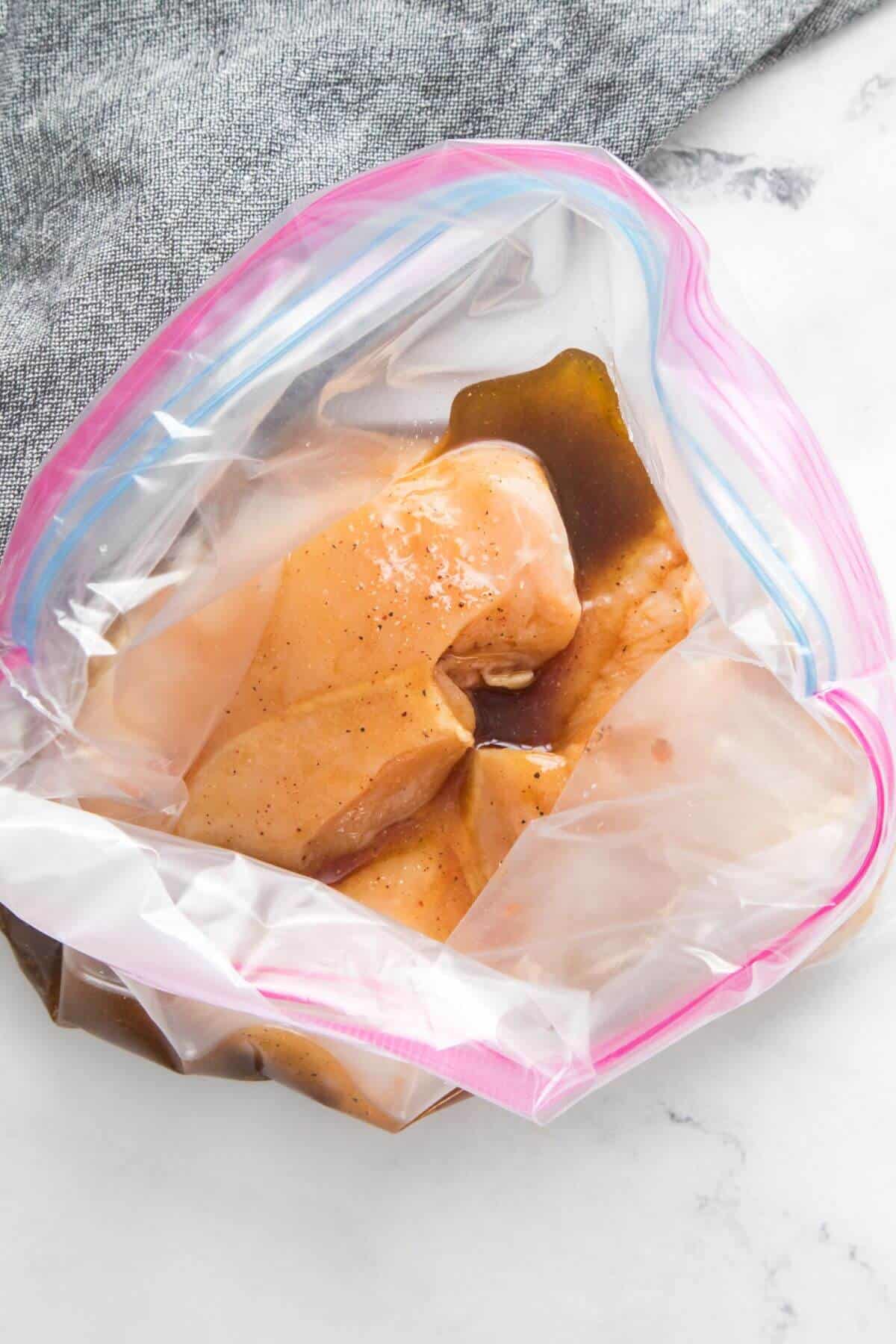 Raw chicken marinating in a sealed plastic bag placed on a white surface with a gray cloth nearby.