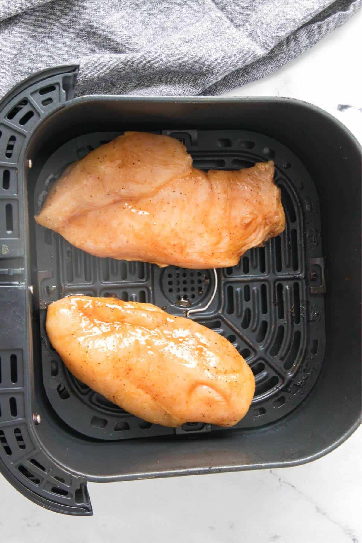 Two raw marinated chicken breasts in an air fryer basket on a marble surface, with a gray cloth nearby.
