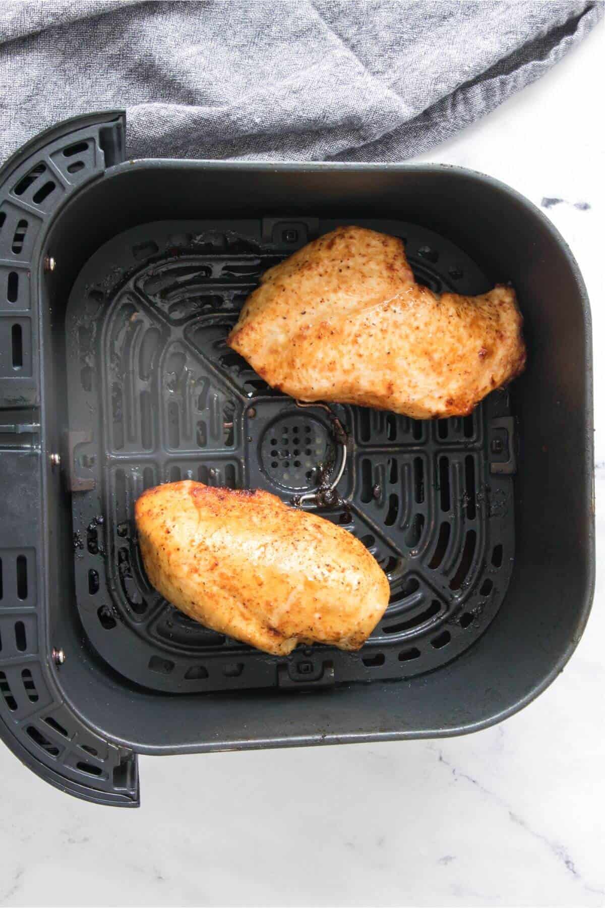 Two cooked chicken breasts in an air fryer basket, set on a white marble surface with a gray cloth nearby.