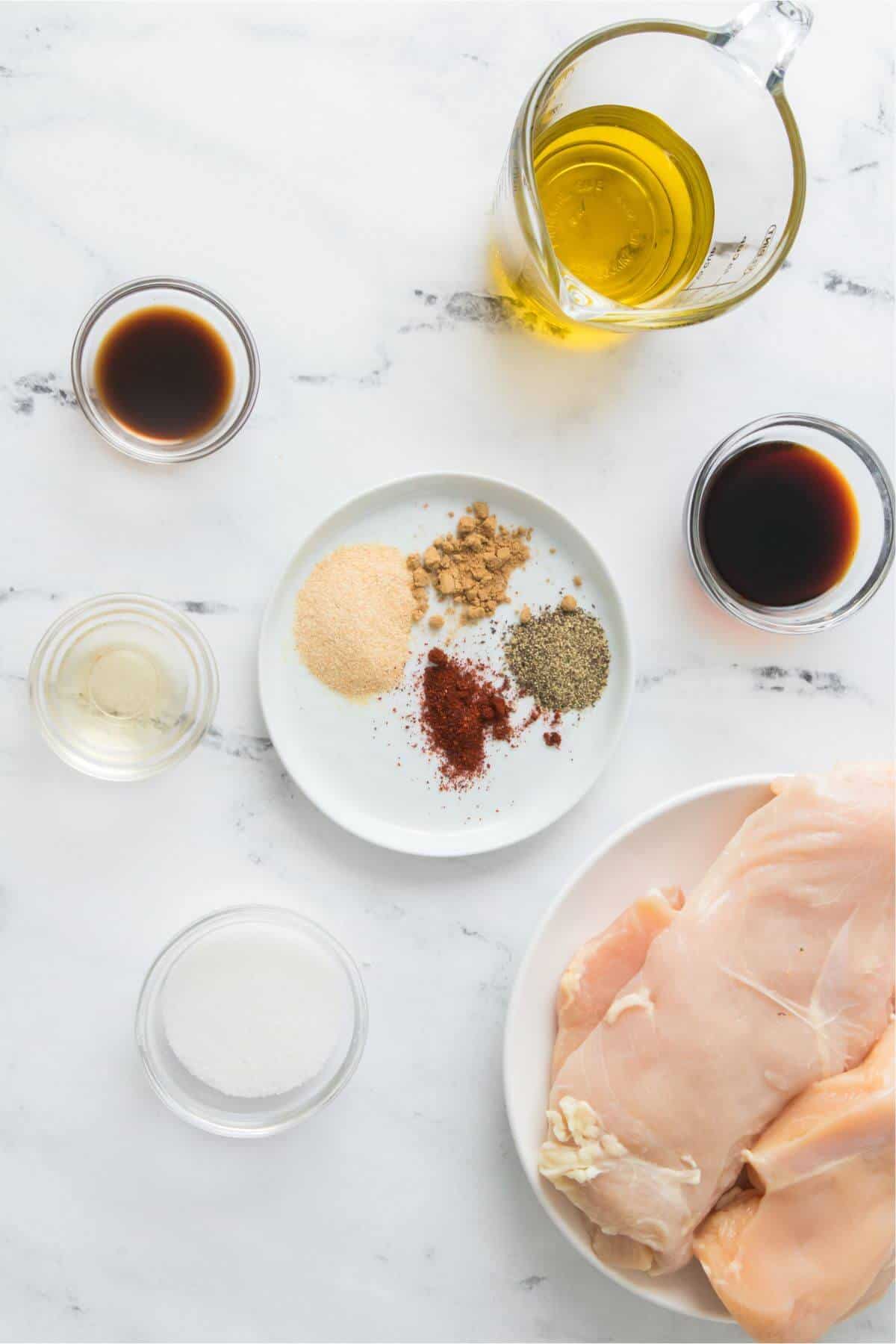 Raw chicken breasts with olive oil, soy sauce, vinegar, sugar, and a plate of spices on a marble surface.