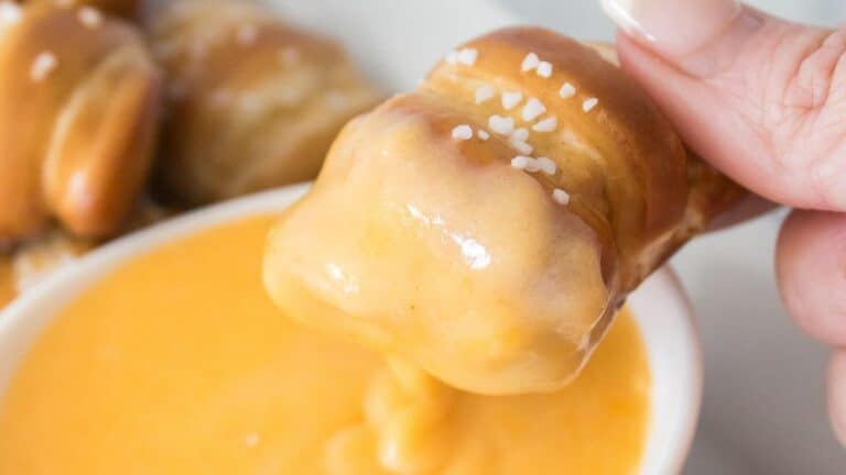 A hand dips a pretzel bite into a bowl of cheese sauce.