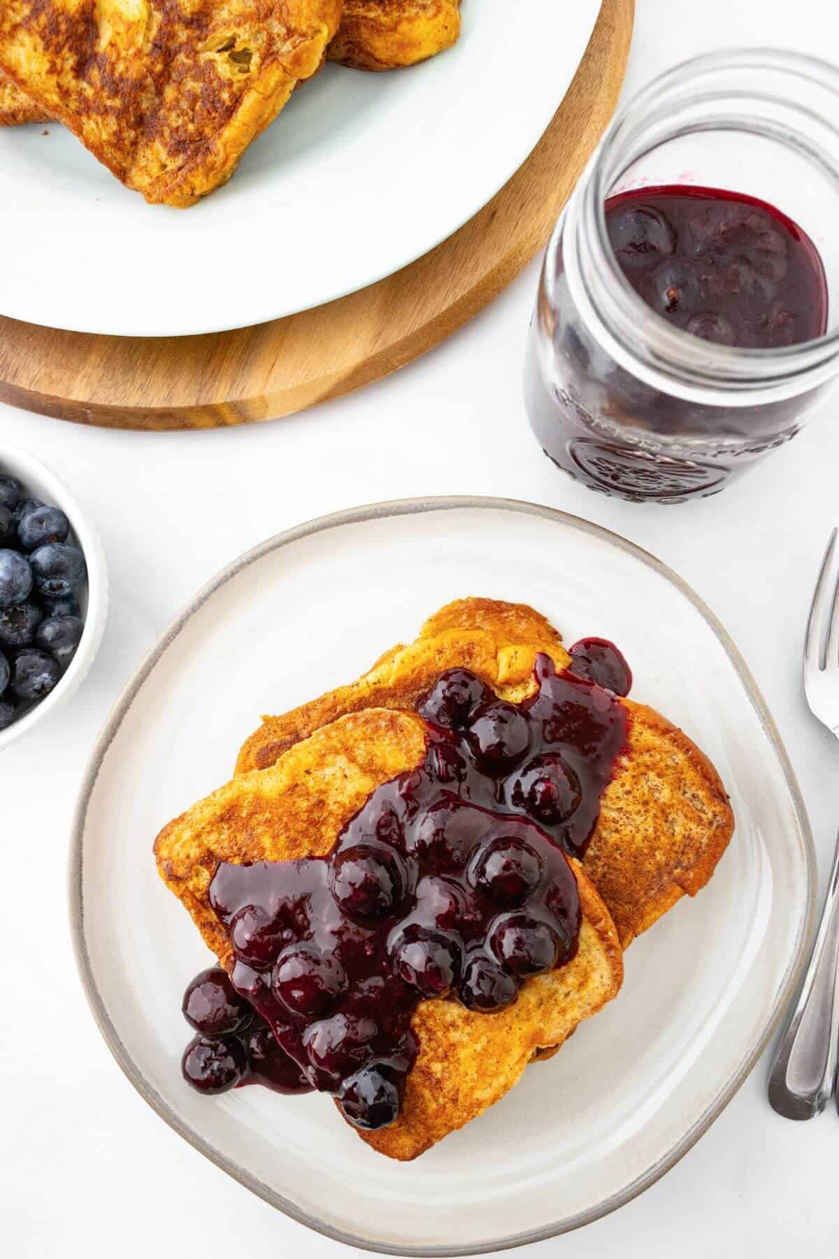 A plate of French toast topped with berry sauce sits on a table. Nearby are a jar of sauce, a bowl of blueberries, a fork, and a spoon. Another plate of French toast is in the background.