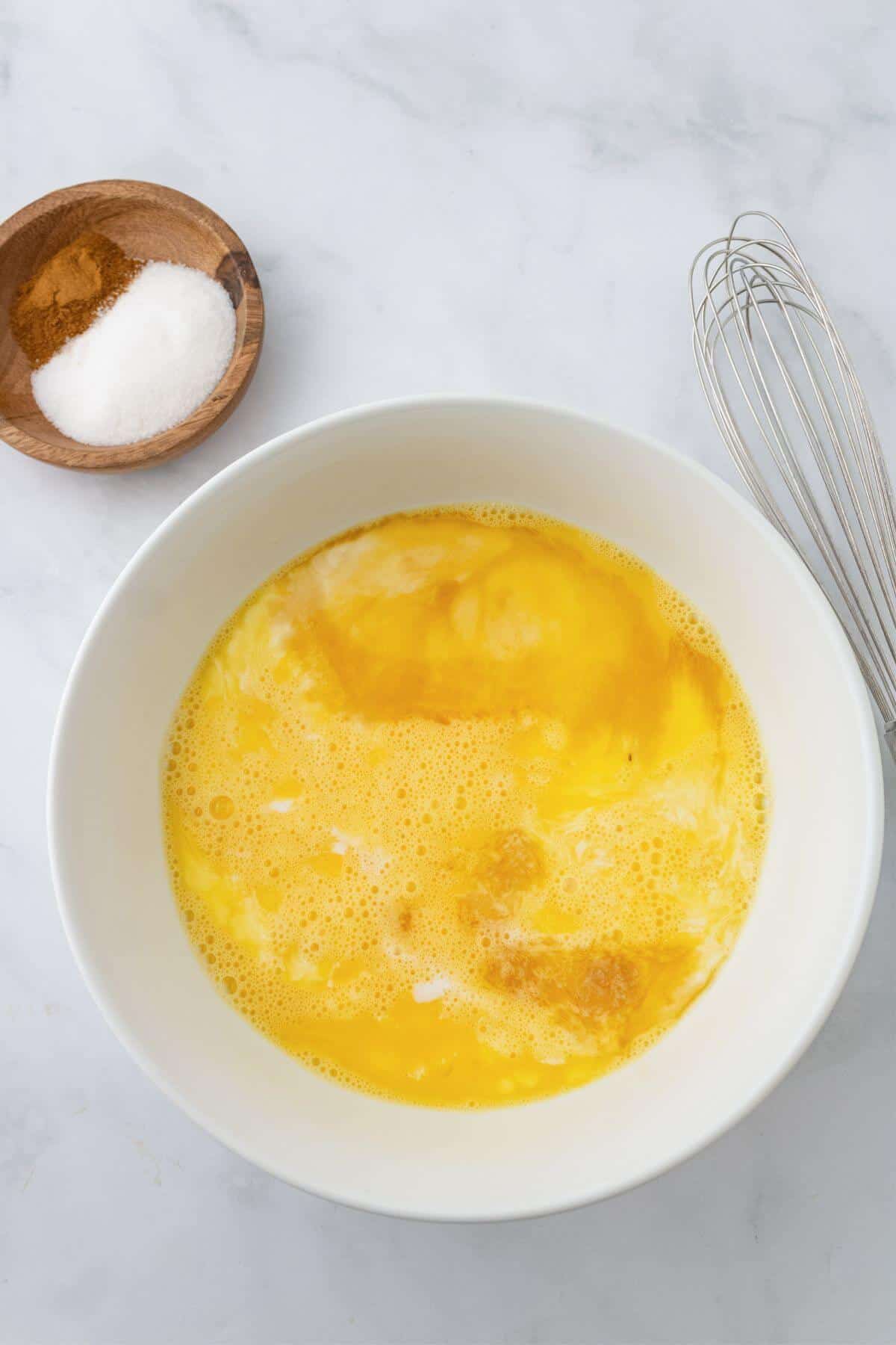 A white bowl contains beaten eggs. Nearby are a whisk and a wooden bowl with salt and cinnamon on a marble surface.