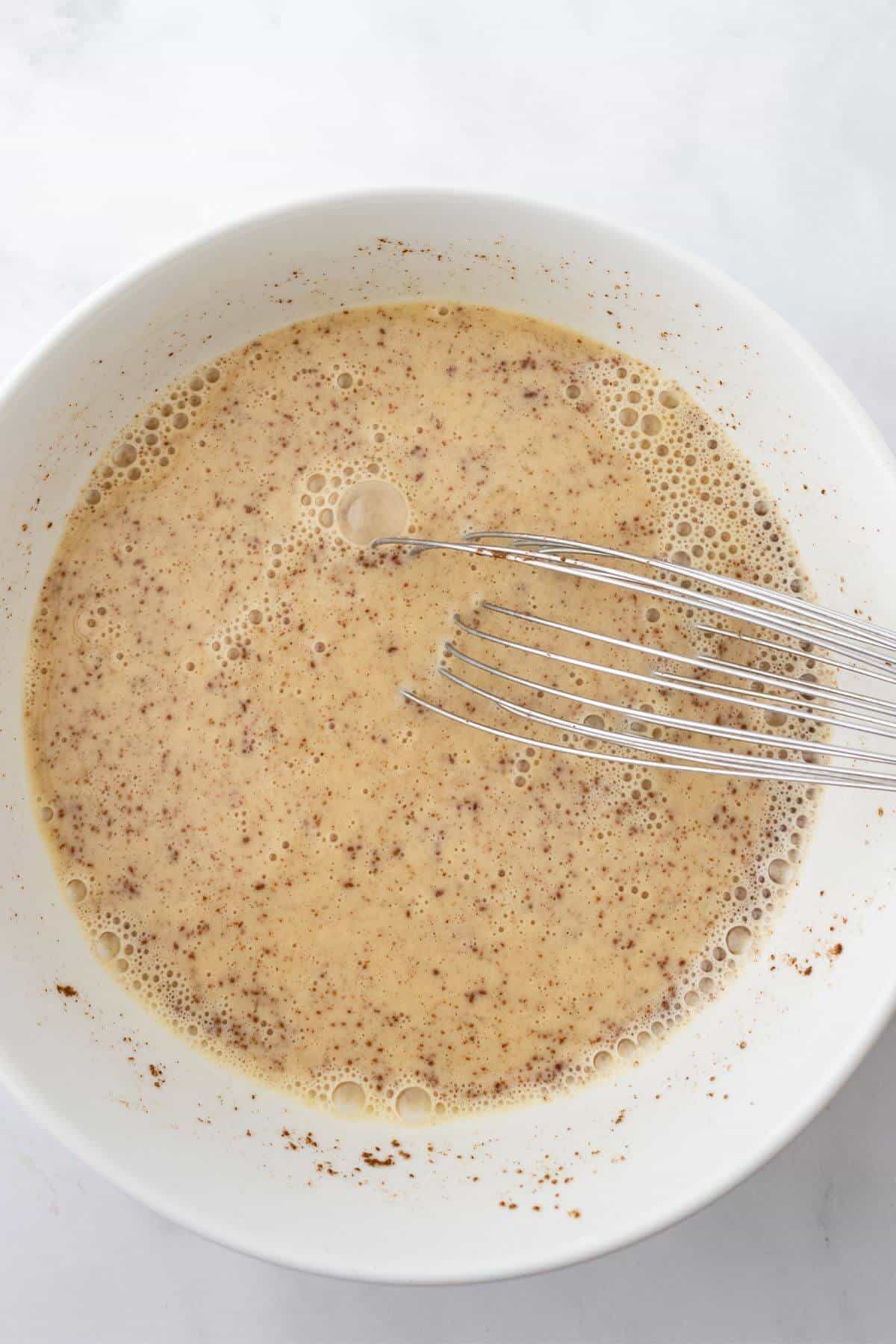 A bowl of whisked eggs and spices with visible bubbles on the surface, and a metal whisk resting inside.