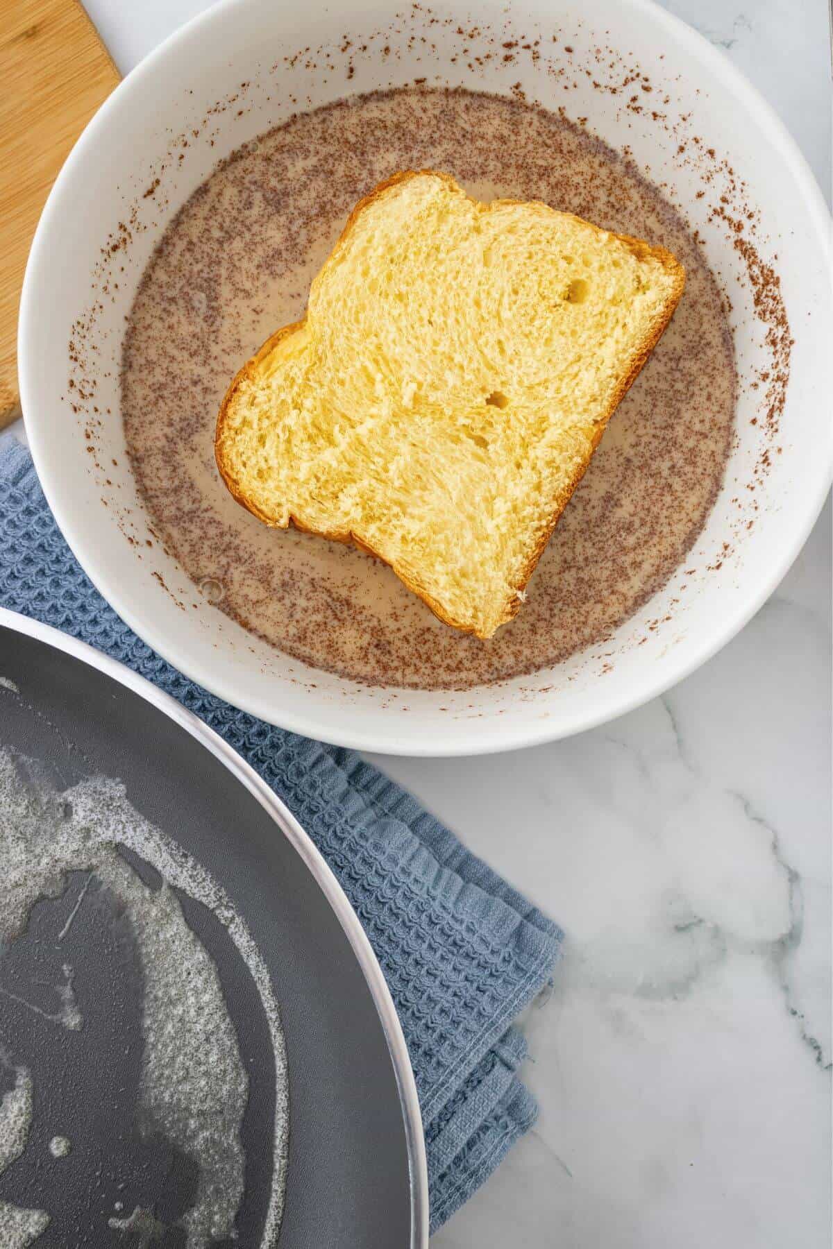 A slice of bread soaking in a mixture within a white bowl, next to a frying pan with melted butter, on a marble surface with a blue cloth.