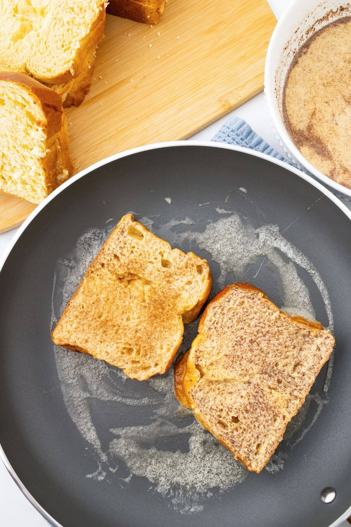 Two slices of French toast cooking in a frying pan, with a bowl of egg mixture and bread slices on a cutting board in the background.