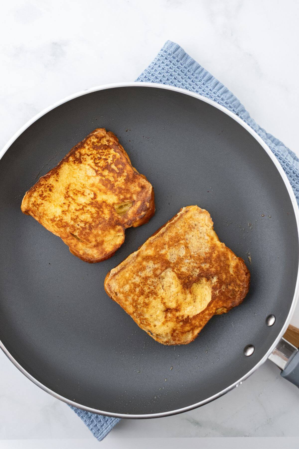 Two slices of French toast cooking in a non-stick frying pan on a marble surface with a blue cloth underneath.