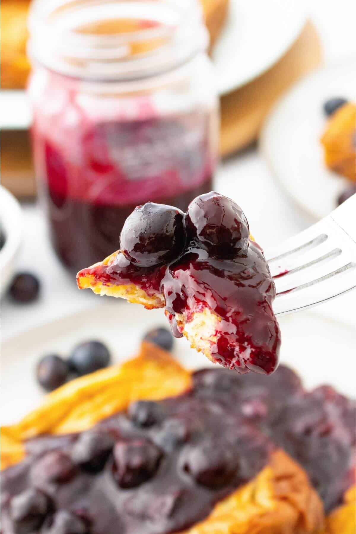 A fork holds a piece of pastry topped with blueberries and purple syrup. A jar and a pastry on a plate are in the background.