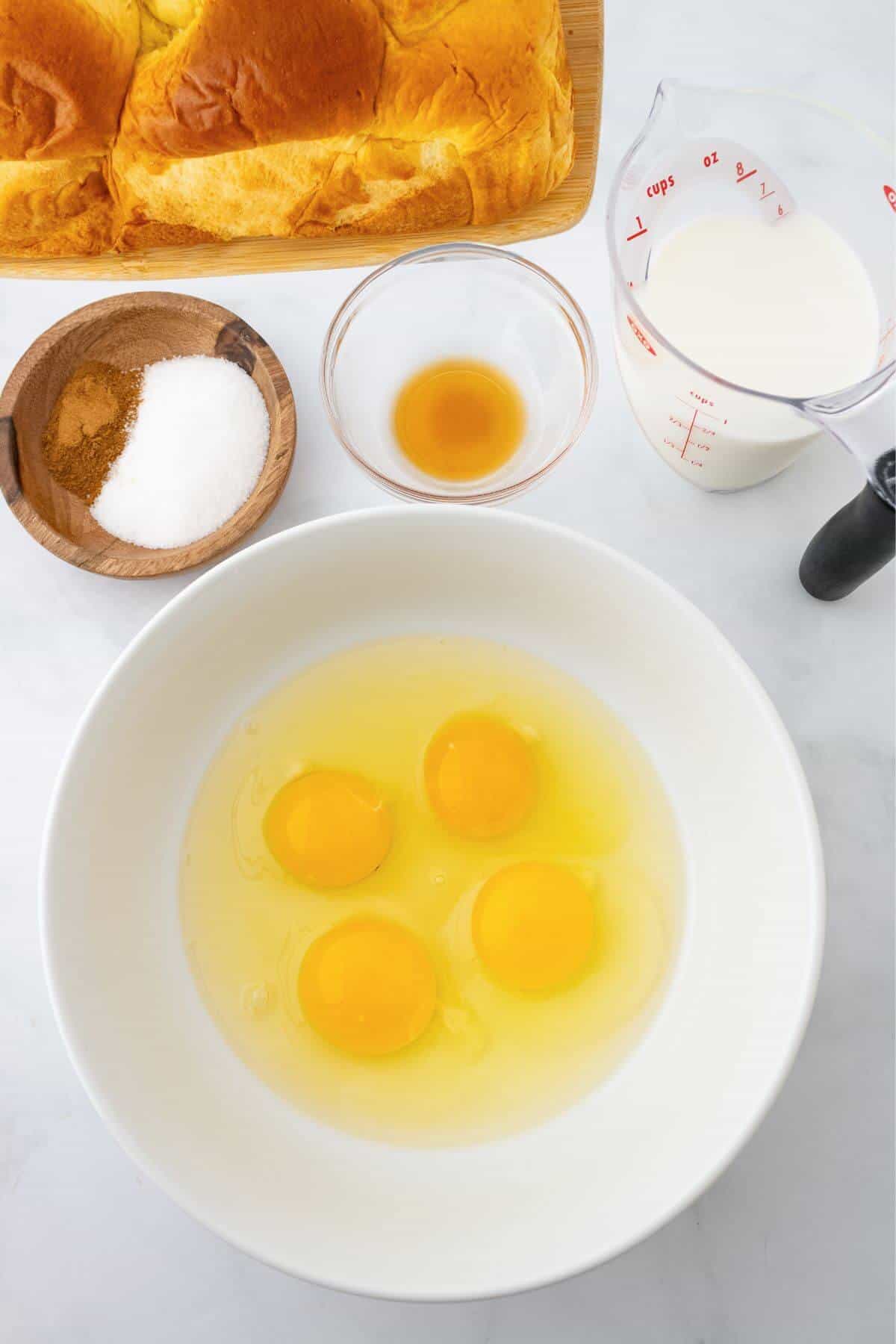 Ingredients for a recipe: three eggs in a bowl, a measuring cup of milk, small bowls of vanilla and sugar, cinnamon, and a loaf of bread.