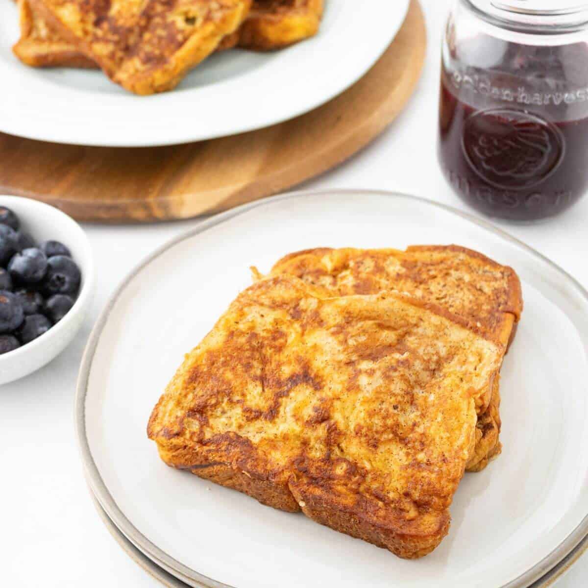 Two slices of French toast on a white plate, with a bowl of blueberries and a jar of dark syrup in the background.