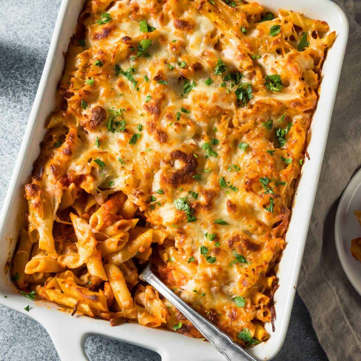 A lazy cook's delight, this baked pasta casserole is topped with melted cheese and garnished with parsley. It's served in a white rectangular dish, complete with a serving spoon on the side for easy scooping.