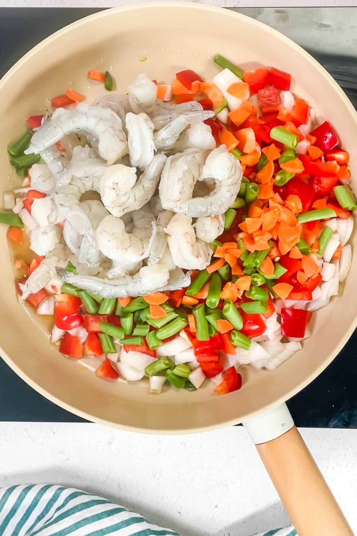 Raw shrimp, chopped green beans, bell peppers, carrots, and onions sizzle in a beige frying pan on a stovetop, perfect for creating keto fried cauliflower rice with shrimp.