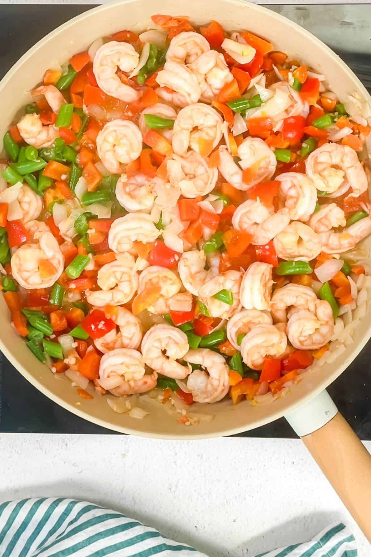 A pan filled with cooked shrimp, diced red and green bell peppers, and onions on a stovetop sizzles with keto fried cauliflower rice. A striped cloth is visible at the bottom.