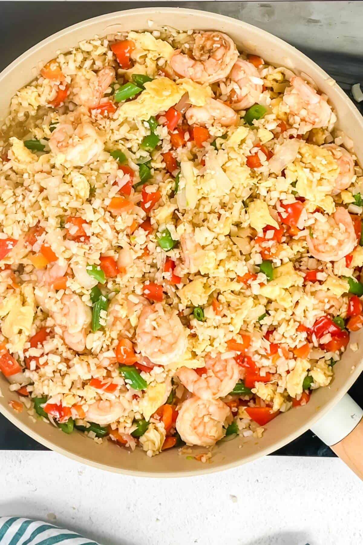 A skillet filled with keto fried cauliflower rice with shrimp, featuring succulent shrimp, fluffy scrambled eggs, crisp green beans, and vibrant diced red bell peppers.