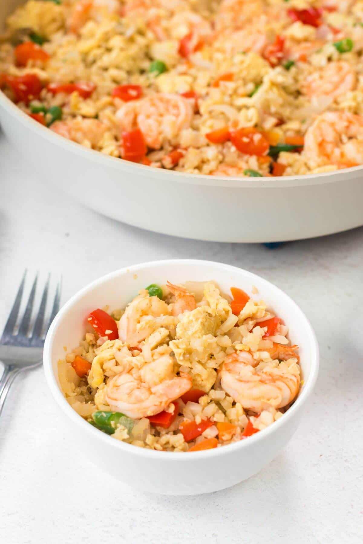 A bowl of shrimp stir-fry with vegetables and keto fried cauliflower rice is placed next to a fork and a larger pan of the same dish.