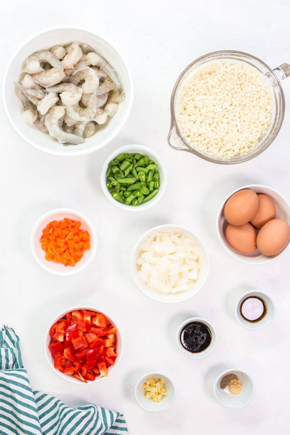 Ingredients for a dish arranged on a table: raw shrimp, rice, green beans, eggs, chopped onions, red bell peppers, garlic, sauces, and seasonings in separate bowls. Striped cloth nearby hints at a delicious keto fried cauliflower rice with shrimp masterpiece waiting to be crafted.