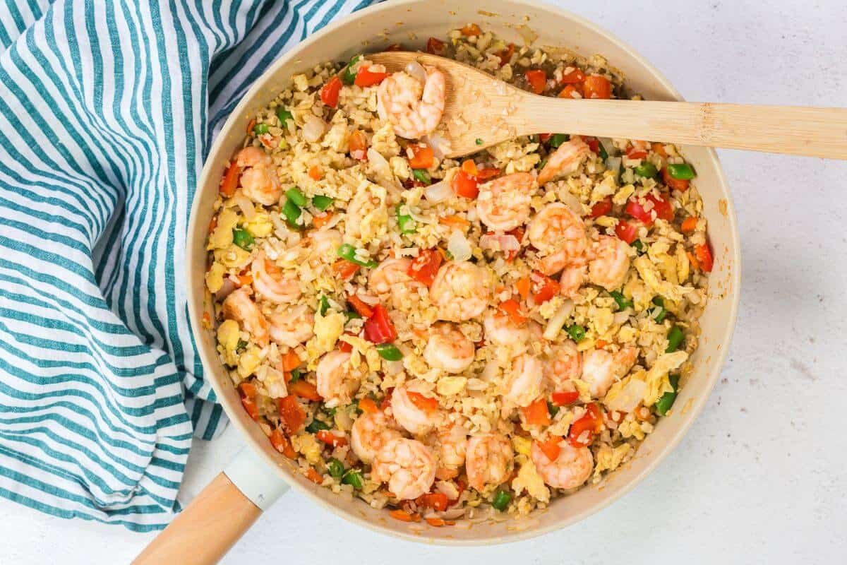 A skillet brimming with keto fried cauliflower rice features shrimp, scrambled eggs, peas, and diced red peppers. A wooden spoon rests beside it on a blue and white striped cloth, ready to serve this delicious low-carb dish.