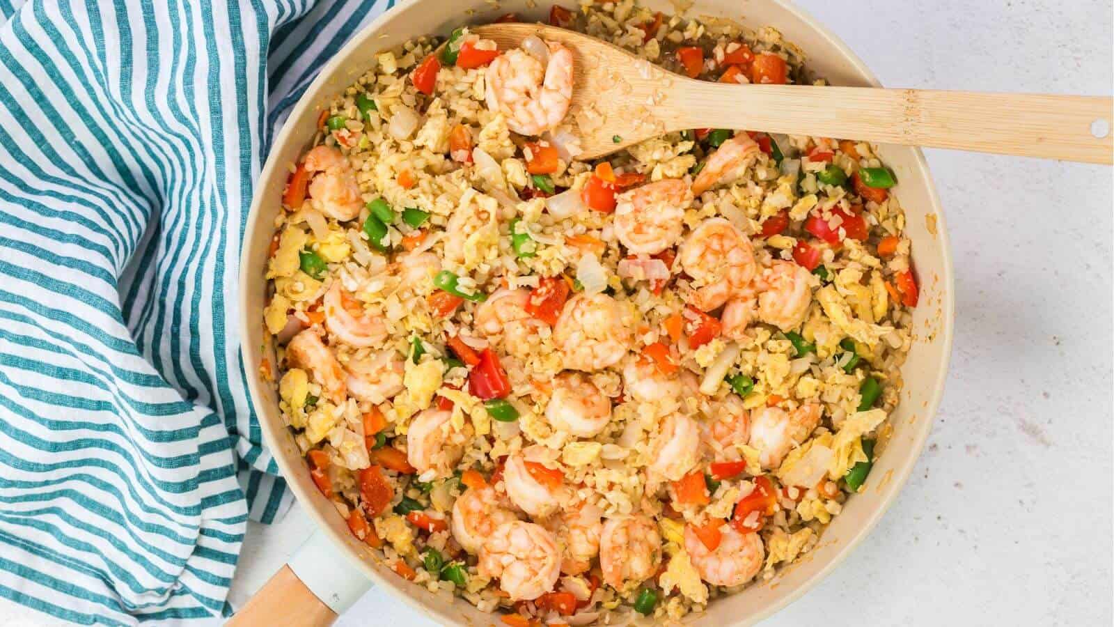 A skillet brimming with keto fried cauliflower rice, shrimp, peas, carrots, and bell peppers is accompanied by scrambled eggs. A wooden spoon rests on the side, while a striped cloth adds charm next to the skillet.