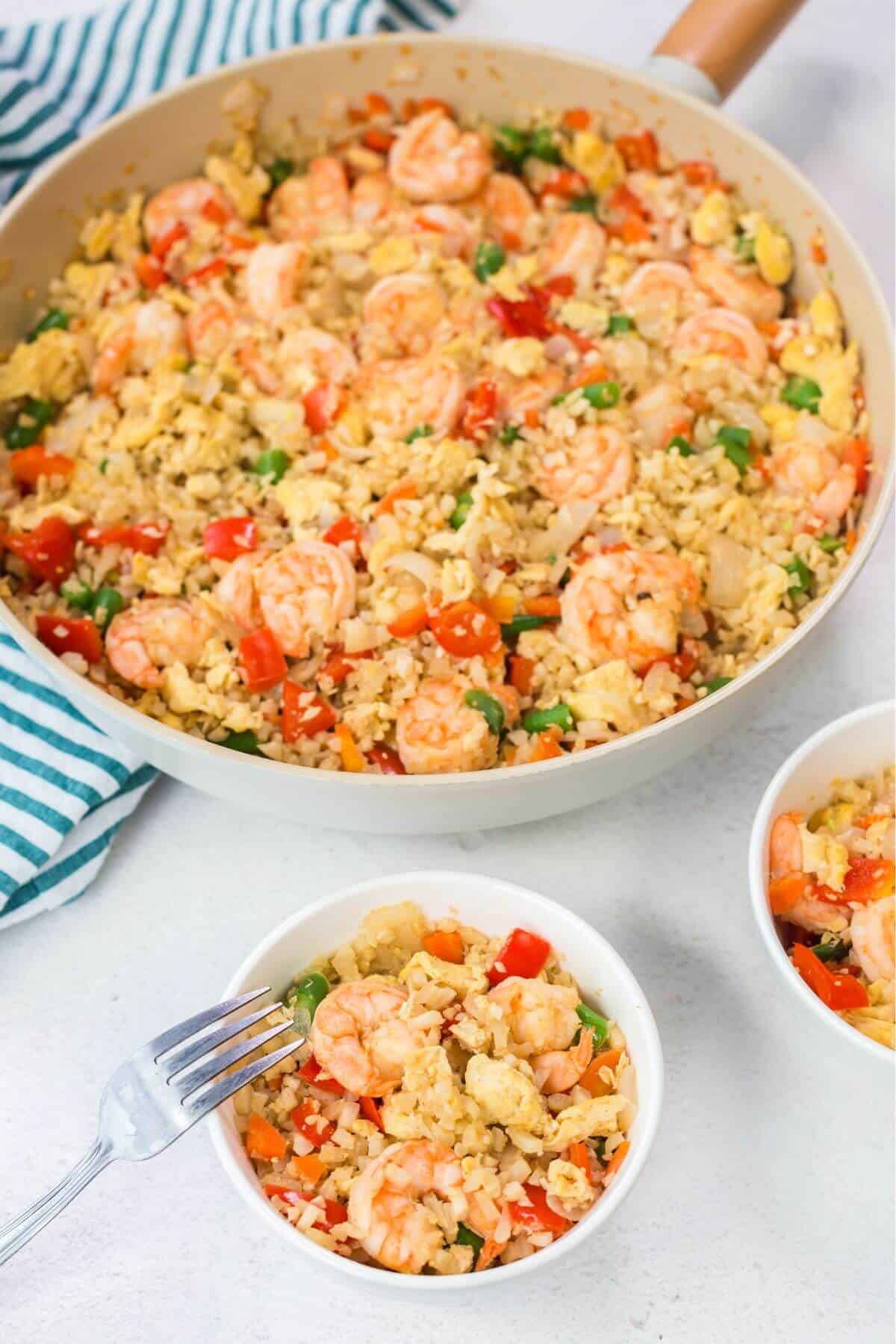 A skillet of keto fried cauliflower rice with shrimp and vegetables sits beside two bowls of the savory dish. A fork rests on one bowl, ready to dive in. A striped cloth is partially visible, adding a touch of style to this delightful meal scene.