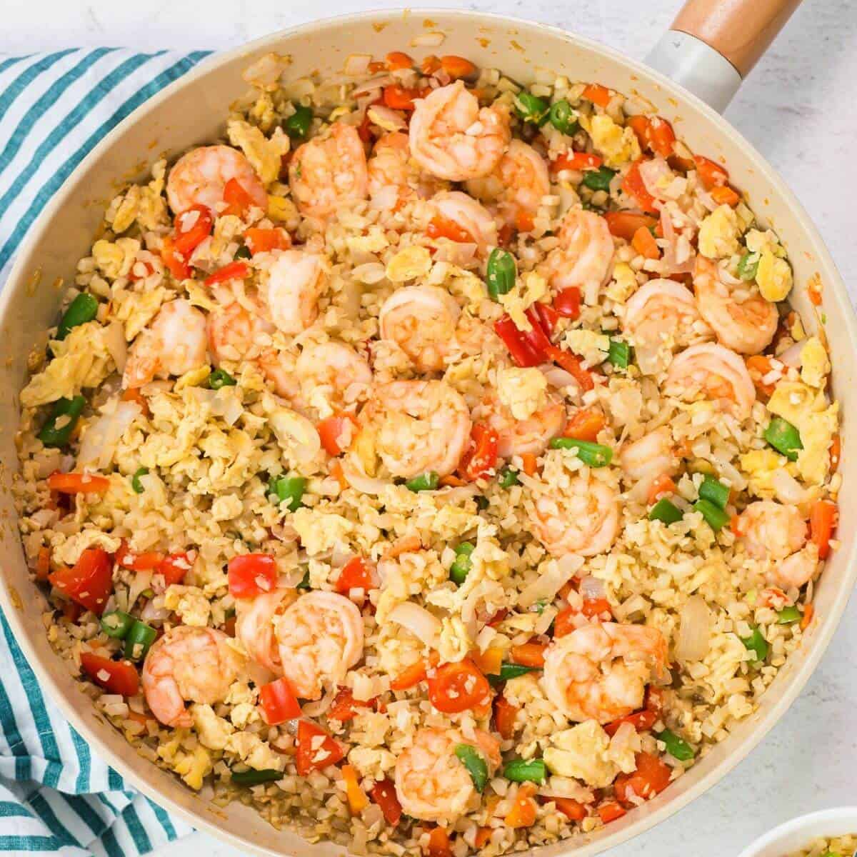 A skillet containing keto fried cauliflower rice with shrimp, featuring vibrant vegetables like bell peppers, onions, and green beans, rests on a striped cloth.