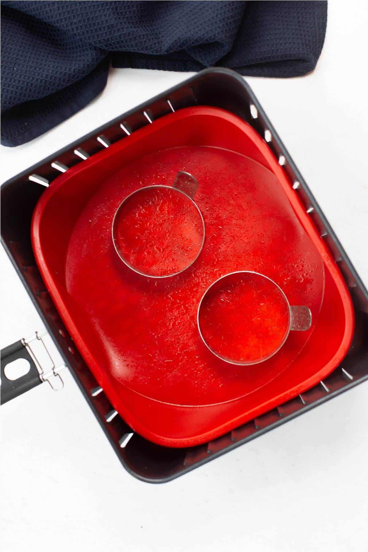 Two round metal cups resembling fried eggs in an air fryer sit in a red shallow tray with a textured surface, nestled inside a black square container.