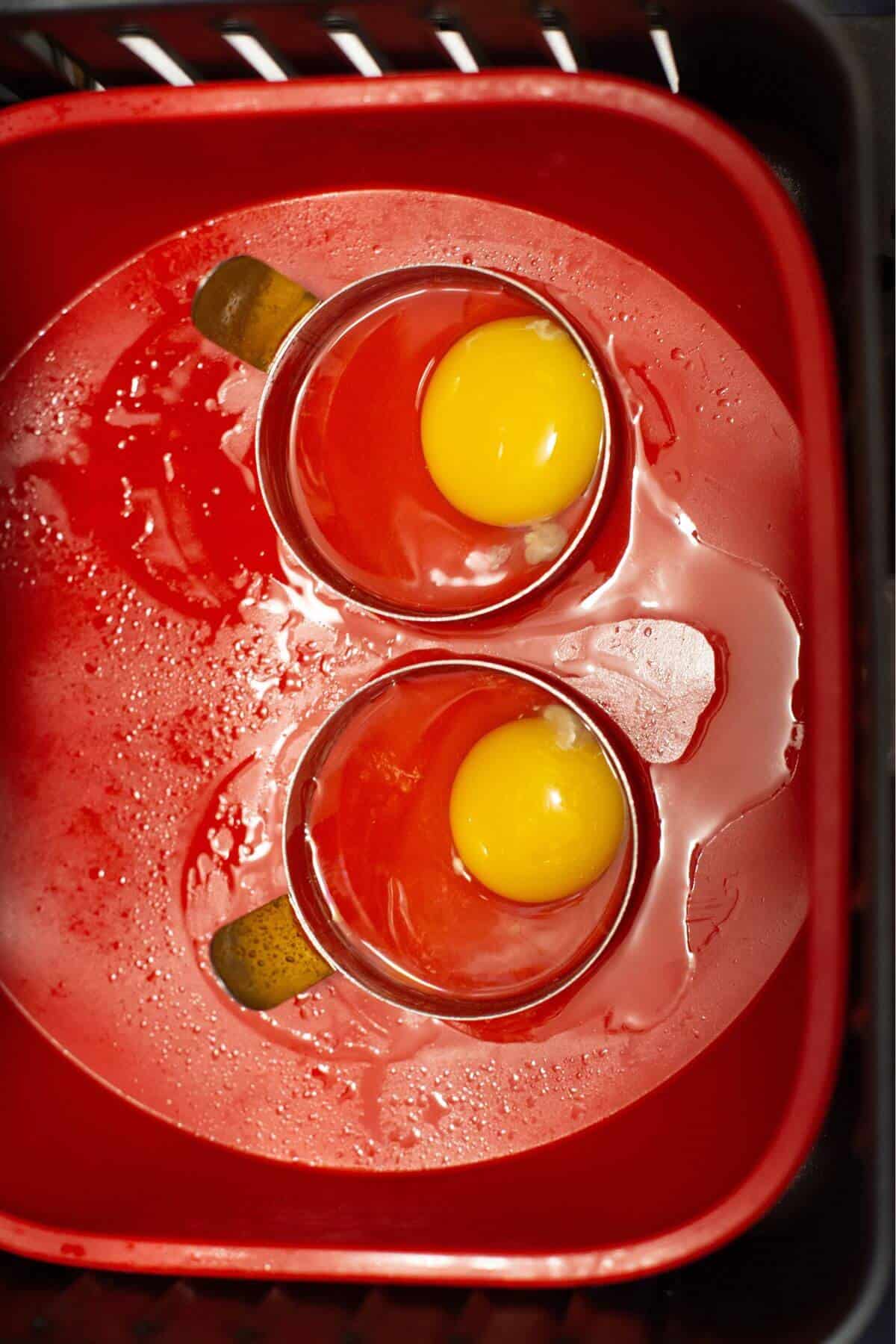 Two cracked eggs with yolks intact sit in mugs, nestled inside a red plastic container with some spilled liquid around them, resembling the hasty preparation of fried eggs in an air fryer.