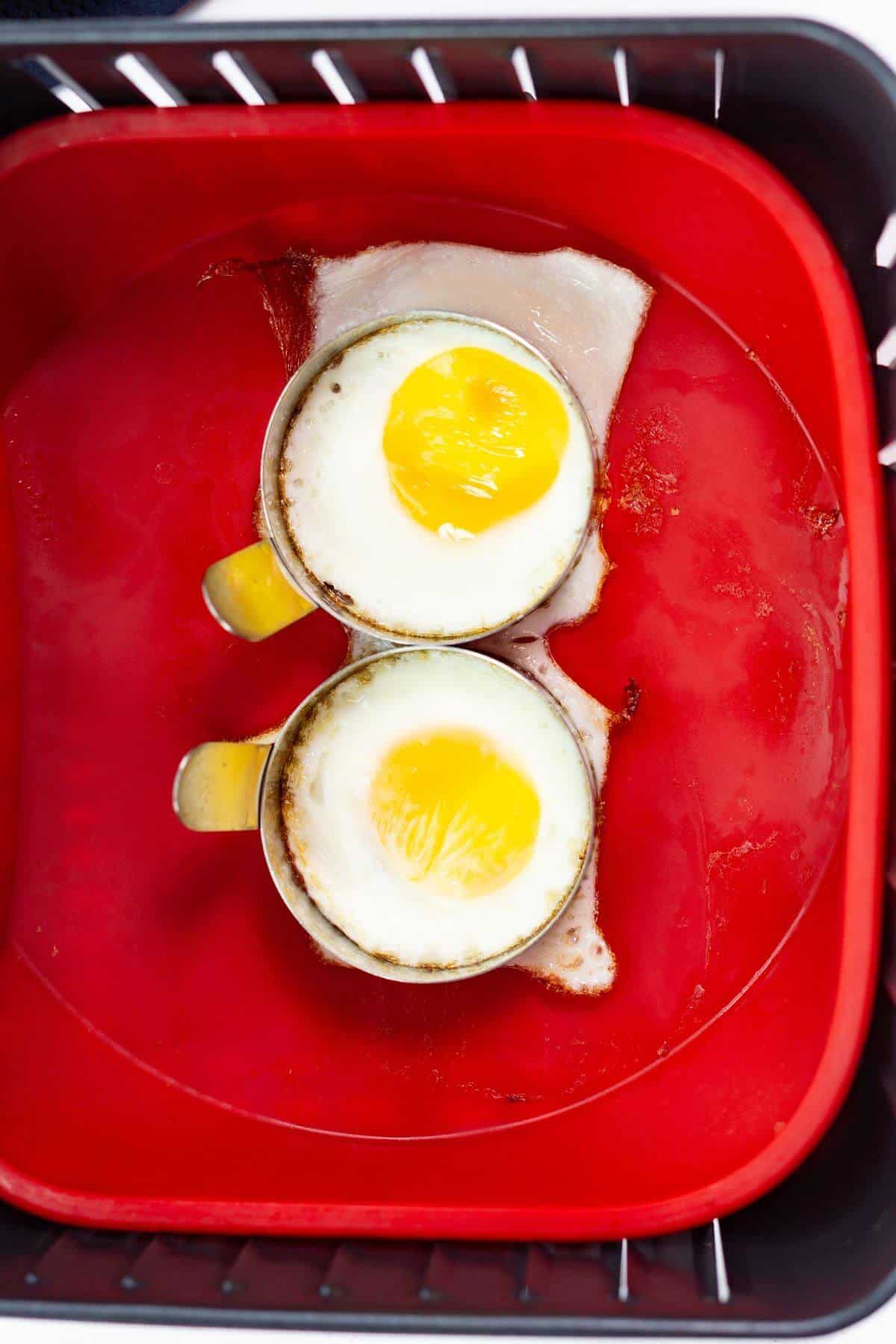 Two fried eggs with sunny-side-up yolks sit in round molds, perfectly fried in the air fryer and placed on sizzling slices of bacon in a red basket.