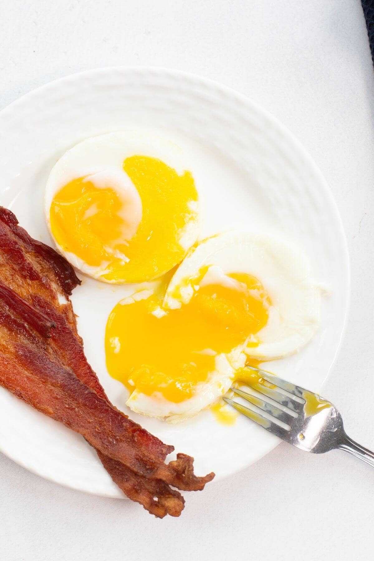 A white plate with two fried eggs crafted in an air fryer, their yolks slightly broken, and two crispy strips of bacon. A fork rests on the plate near the eggs.