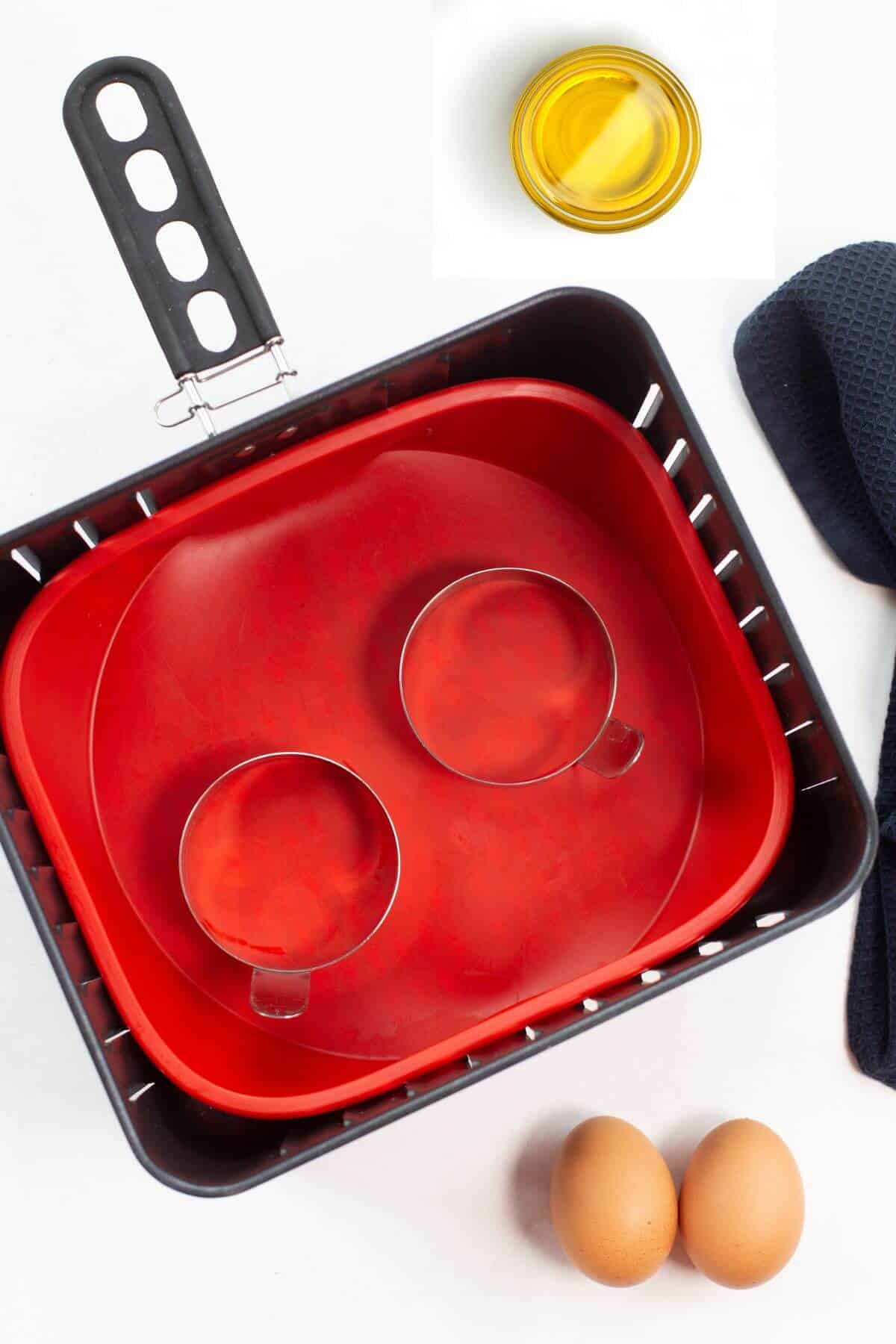 Air fryer basket with a red liner, two metal egg rings ready for frying eggs in the air fryer, a dish of oil, and two eggs on a white surface with a blue cloth nearby.