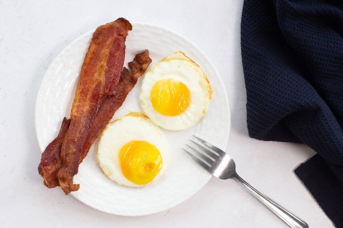 A plate with two perfectly cooked fried eggs from the air fryer and two strips of crispy bacon, accompanied by a fork and dark napkin.