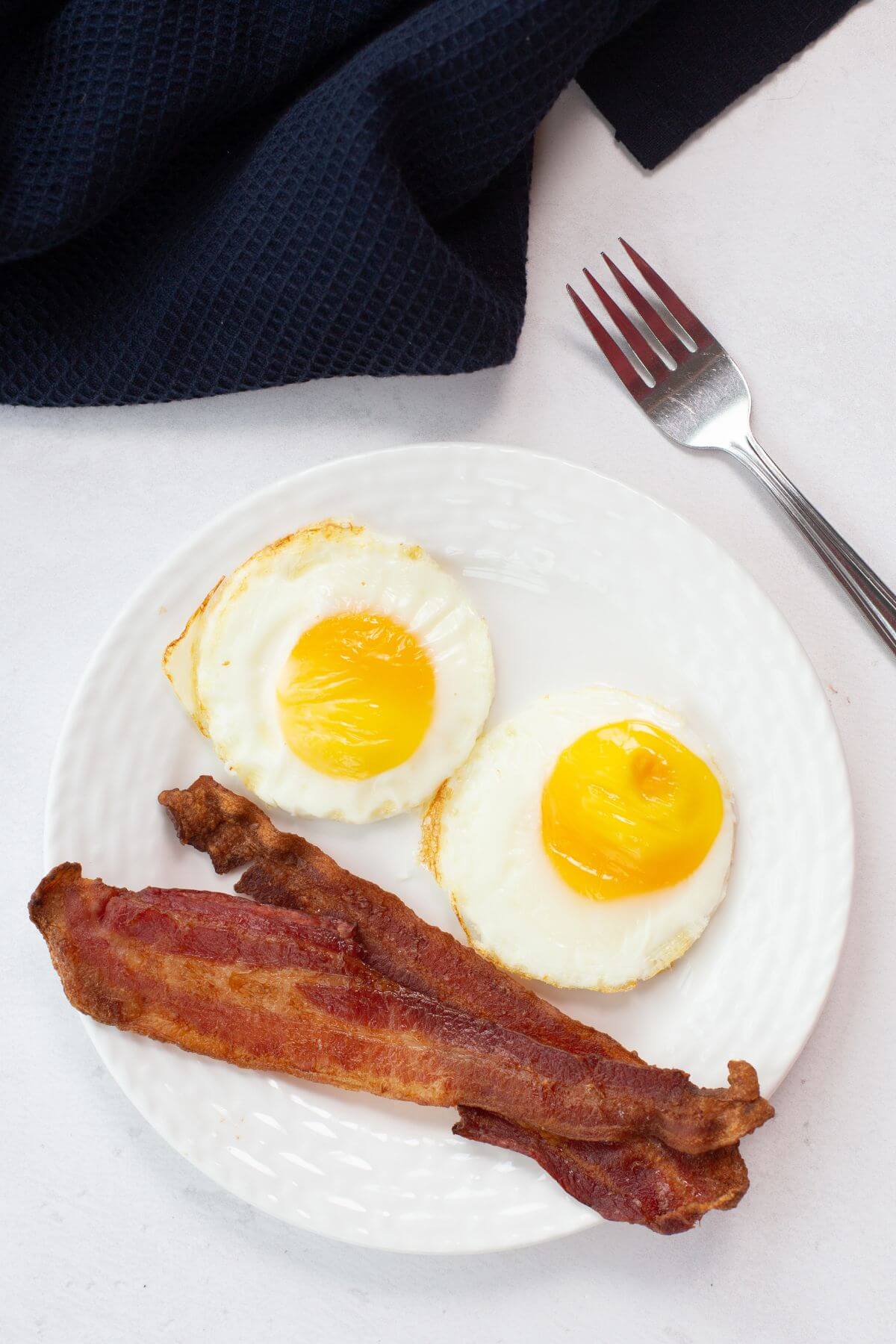 A plate with two sunny-side-up eggs, perfectly resembling those fried in an air fryer, alongside two strips of crispy bacon. A fork rests nearby on a white surface.