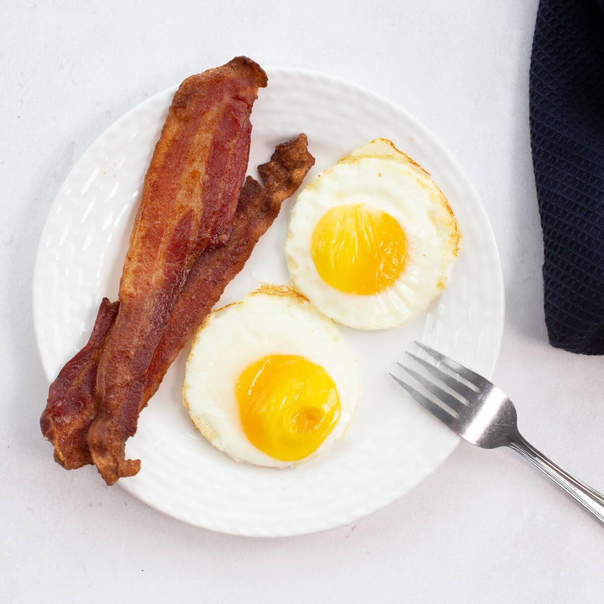A plate featuring two fried eggs, possibly cooked to perfection in an air fryer, and two strips of crispy bacon, accompanied by a fork on a white surface.