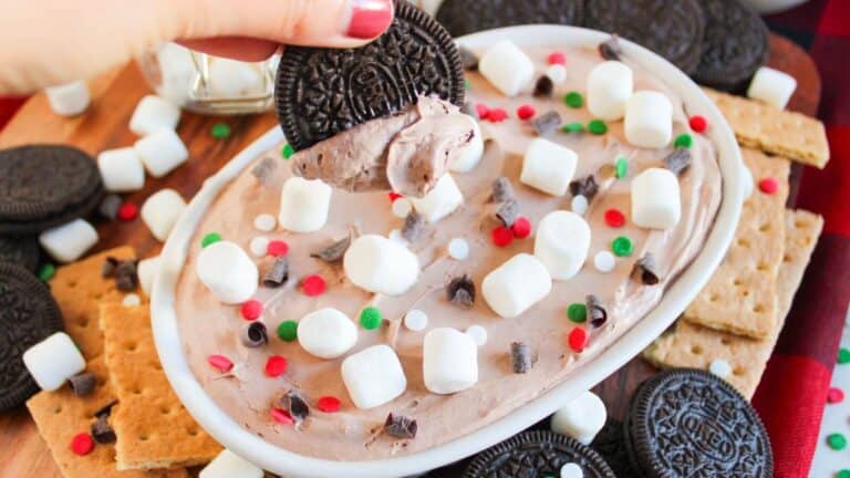 A hand holds a cookie dipped in creamy chocolate spread topped with marshmallows, chocolate chips, and red and green sprinkles. Surrounded by cookies and graham crackers on a platter.