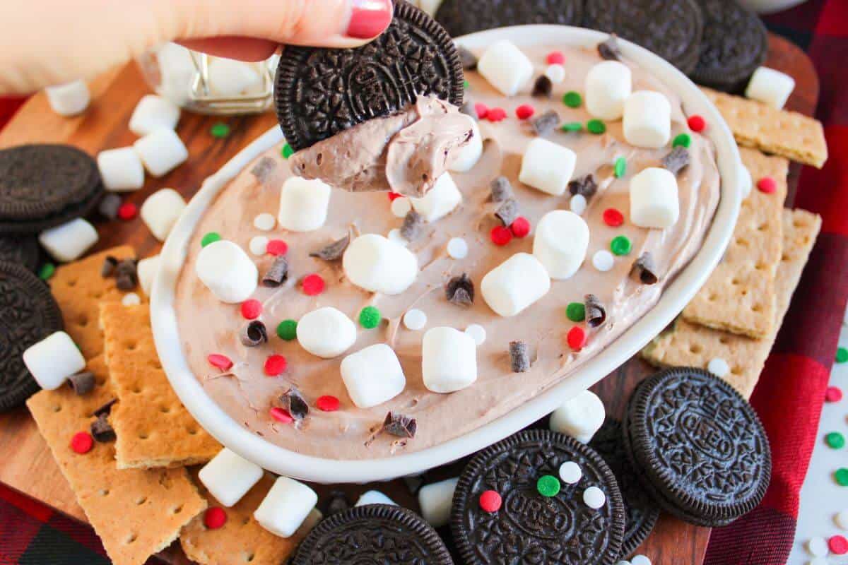 A hand dips a chocolate cookie into a bowl of creamy dip topped with marshmallows and sprinkles, surrounded by more cookies and graham crackers on a wooden tray.