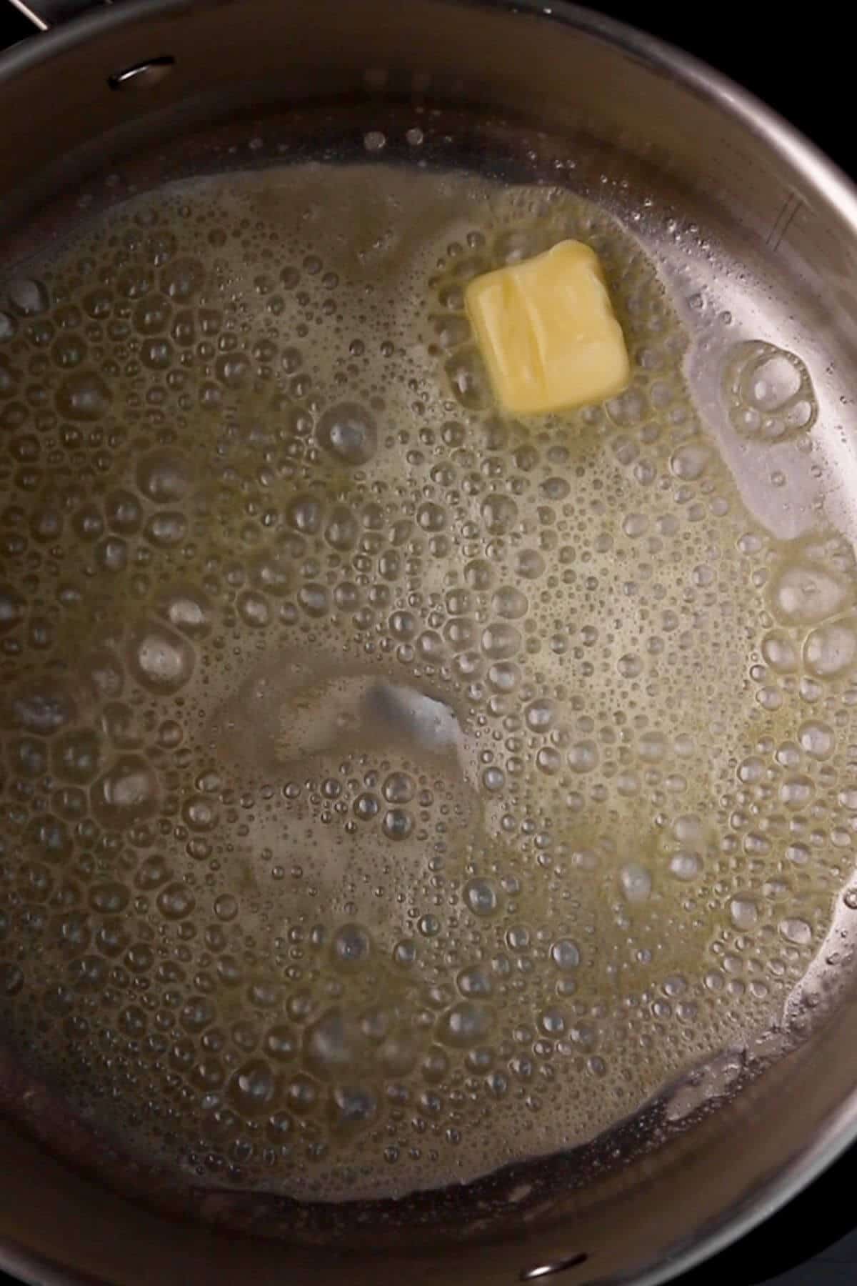 Close-up of a melting butter cube in a stainless steel pan with bubbling liquid.