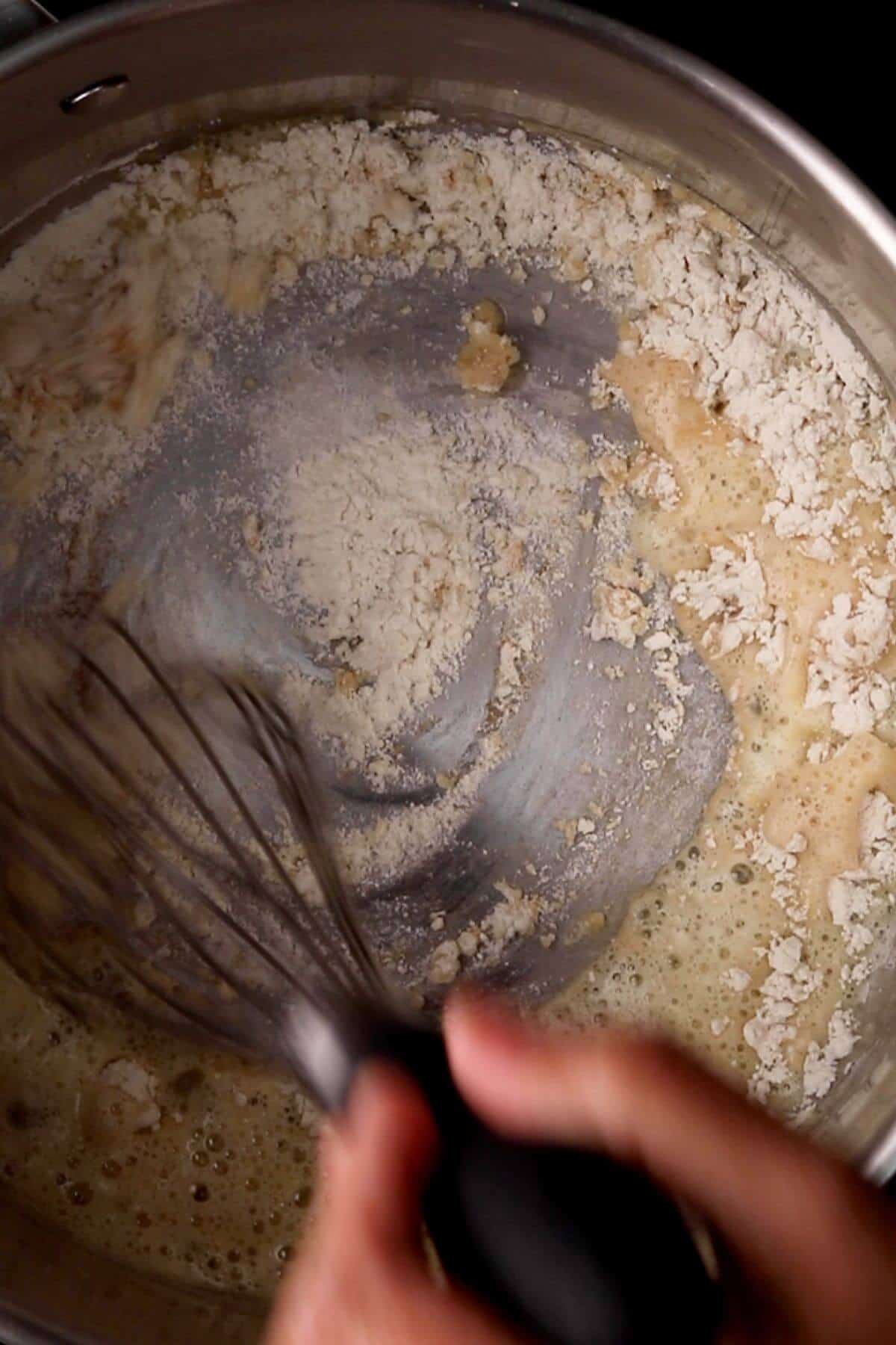 Close-up of a person whisking flour into a pot with liquid, creating a roux.