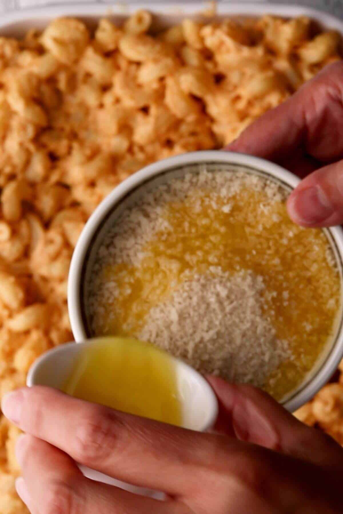 Hands holding a bowl of melted butter and crumbs over a dish of baked macaroni and cheese.