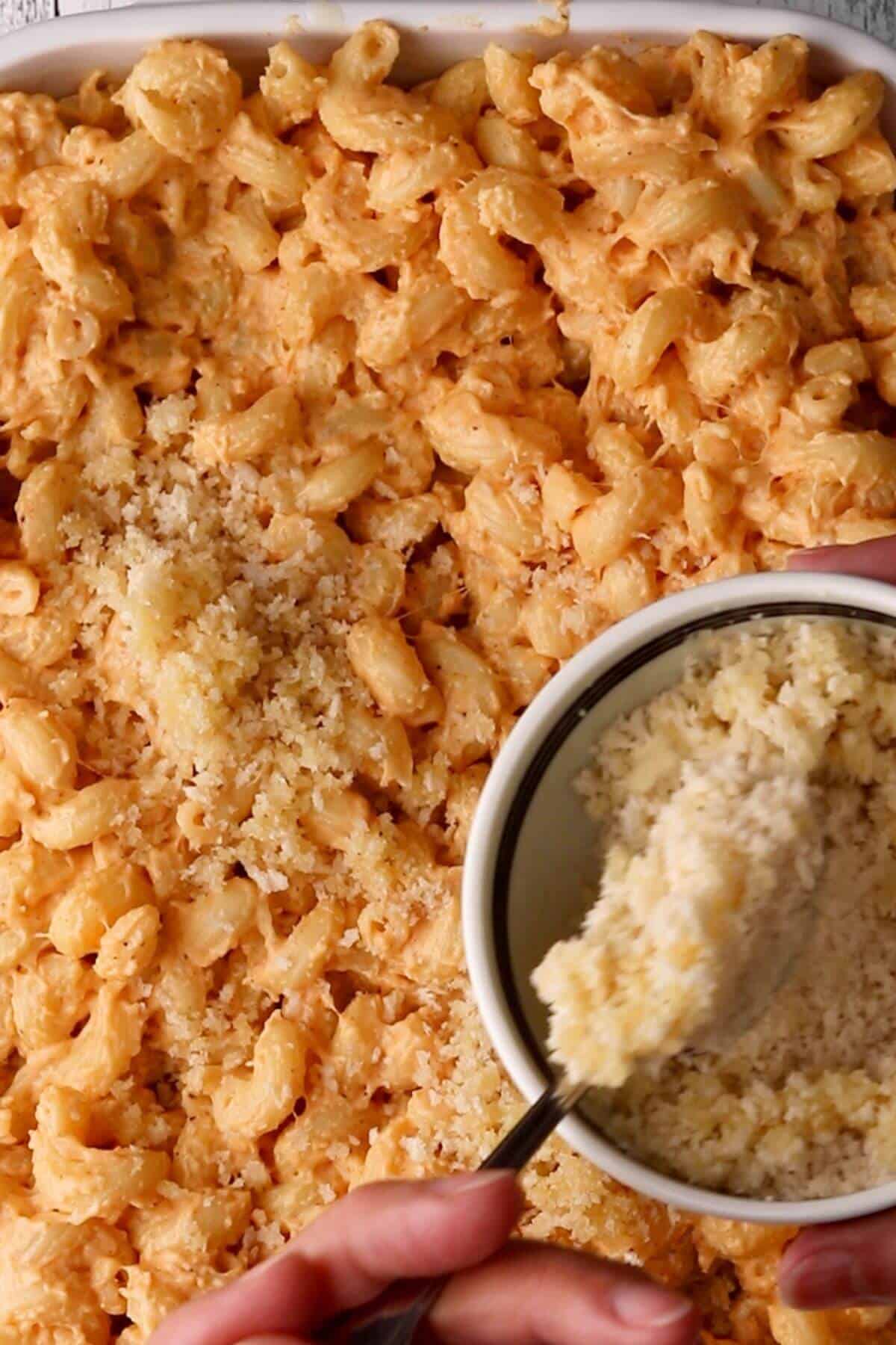 Hands sprinkling breadcrumbs over macaroni and cheese in a baking dish.