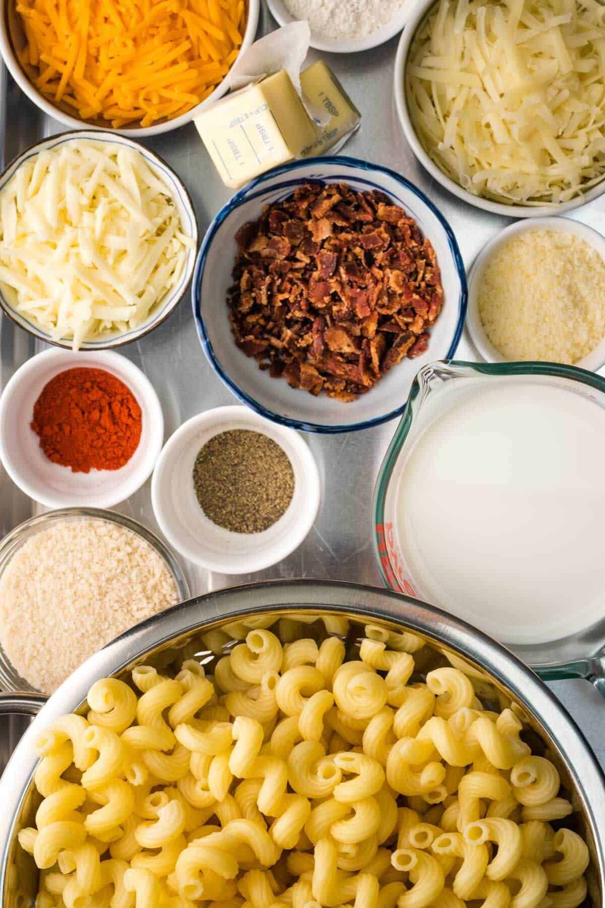 Ingredients for macaroni and cheese are arranged on a table, including pasta, milk, bacon, shredded cheese, butter, and various spices in bowls.