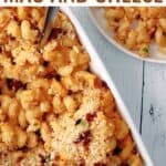 Close-up of a baking dish filled with mac and cheese topped with crispy breadcrumbs and bacon bits, with a serving spoon, placed on a light wooden surface. A small serving is in the background.