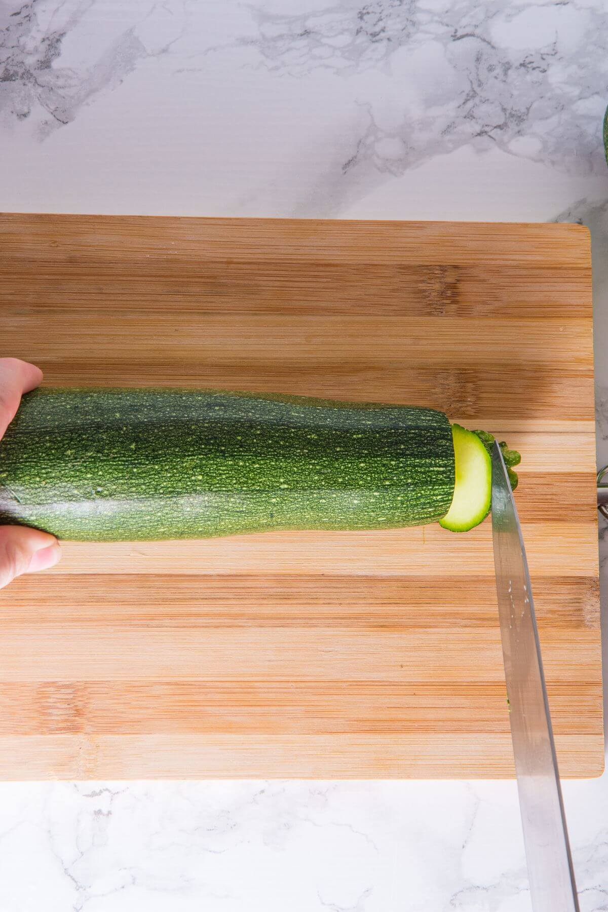 A person expertly slices a zucchini on a wooden cutting board, preparing it for a delicious roasted zucchini dish.