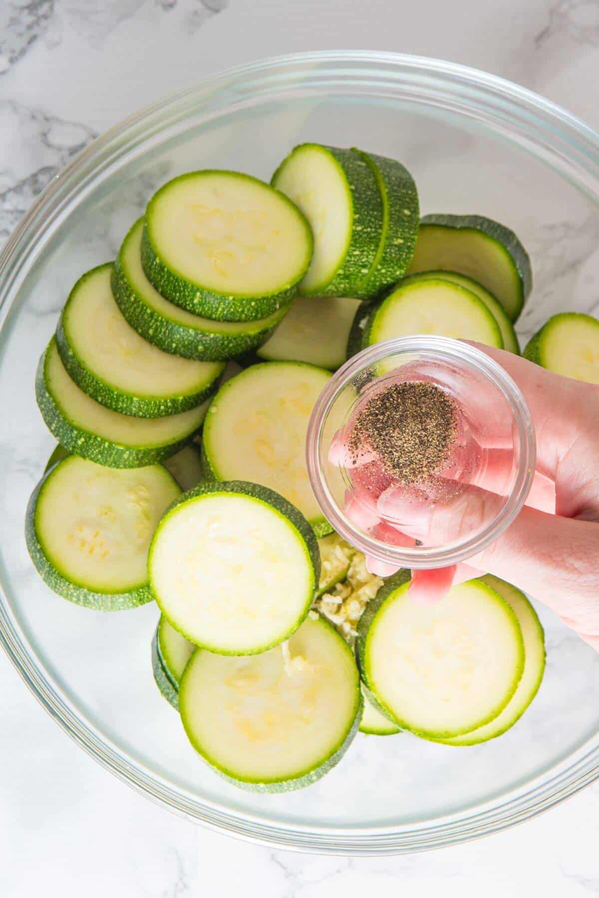 A hand holds a small bowl of ground pepper above a glass bowl filled with sliced zucchini, ready to be transformed into savory roasted zucchini.