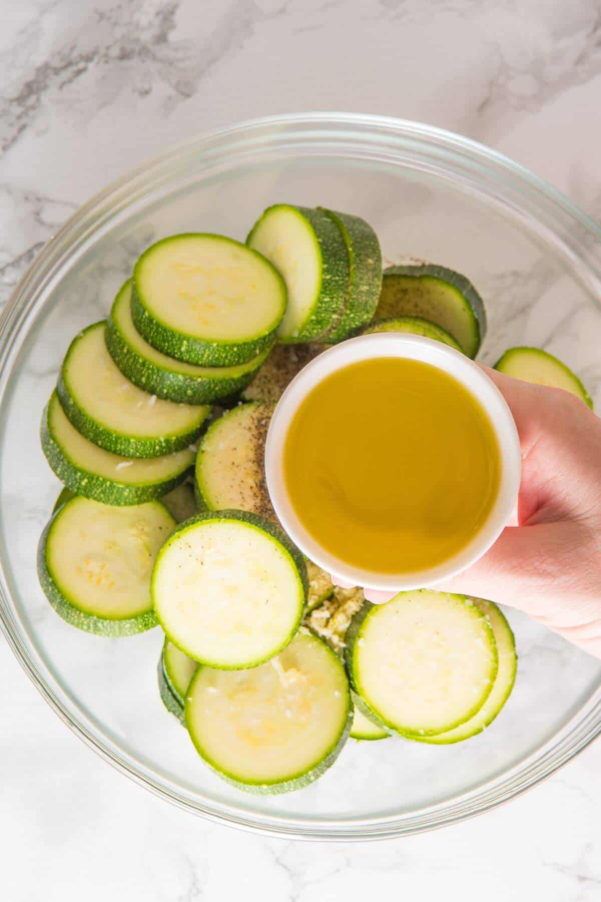 A hand holds a small bowl of olive oil above a glass bowl filled with sliced zucchini on a marble surface, ready to be transformed into delicious roasted zucchini.