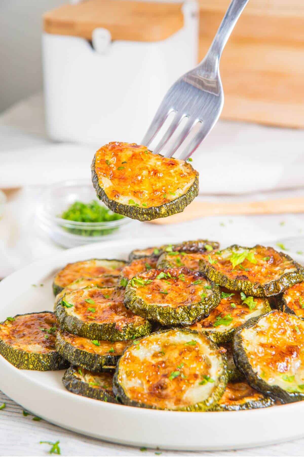A fork lifts a slice of roasted zucchini with melted cheese from a plate stacked with similar slices. A jar and a small dish of herbs are in the background.