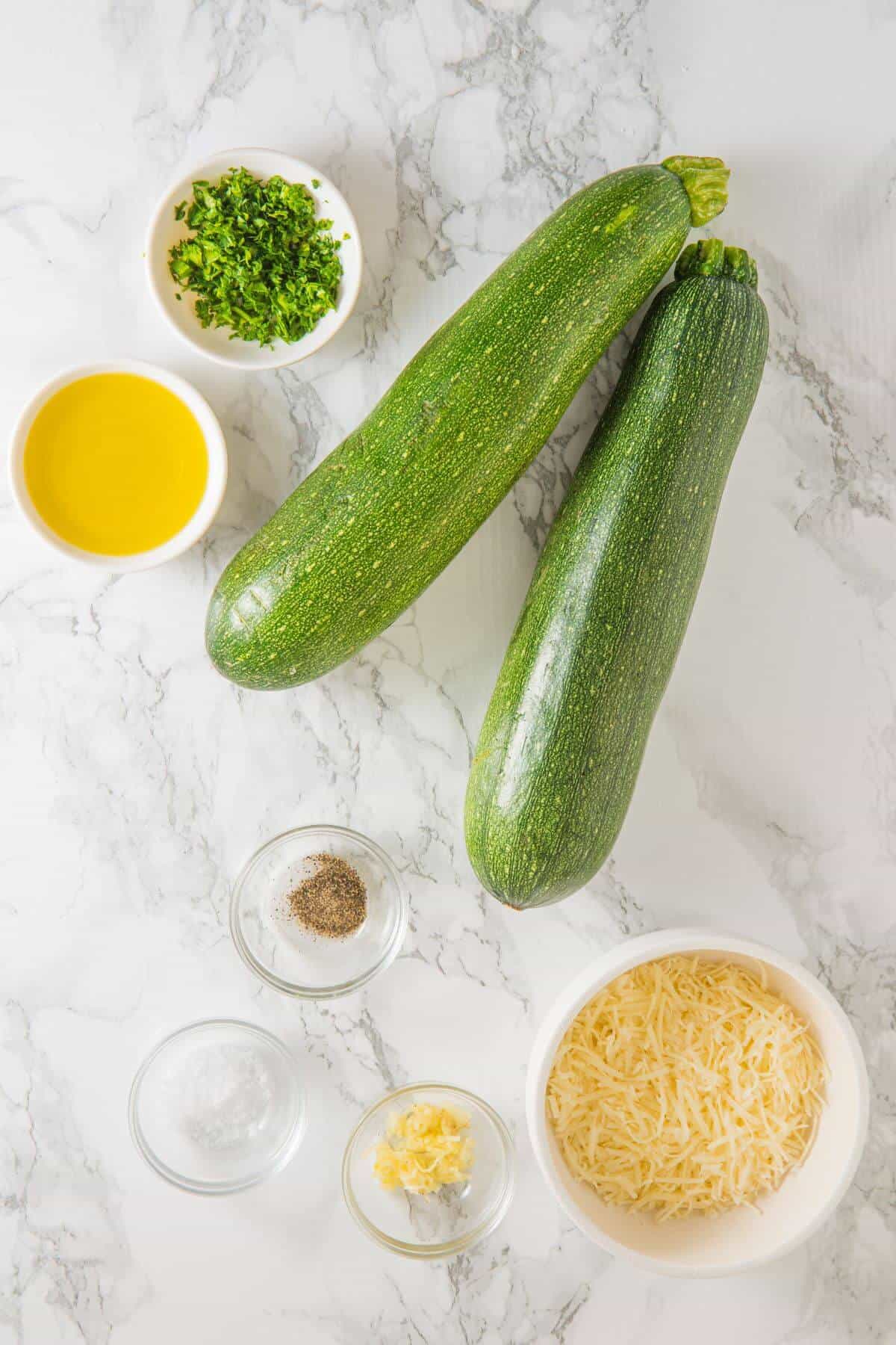 Two zucchinis rest on a marble surface, ready to be transformed into roasted zucchini. They're surrounded by bowls filled with olive oil, chopped herbs, grated cheese, minced garlic, salt, and pepper.
