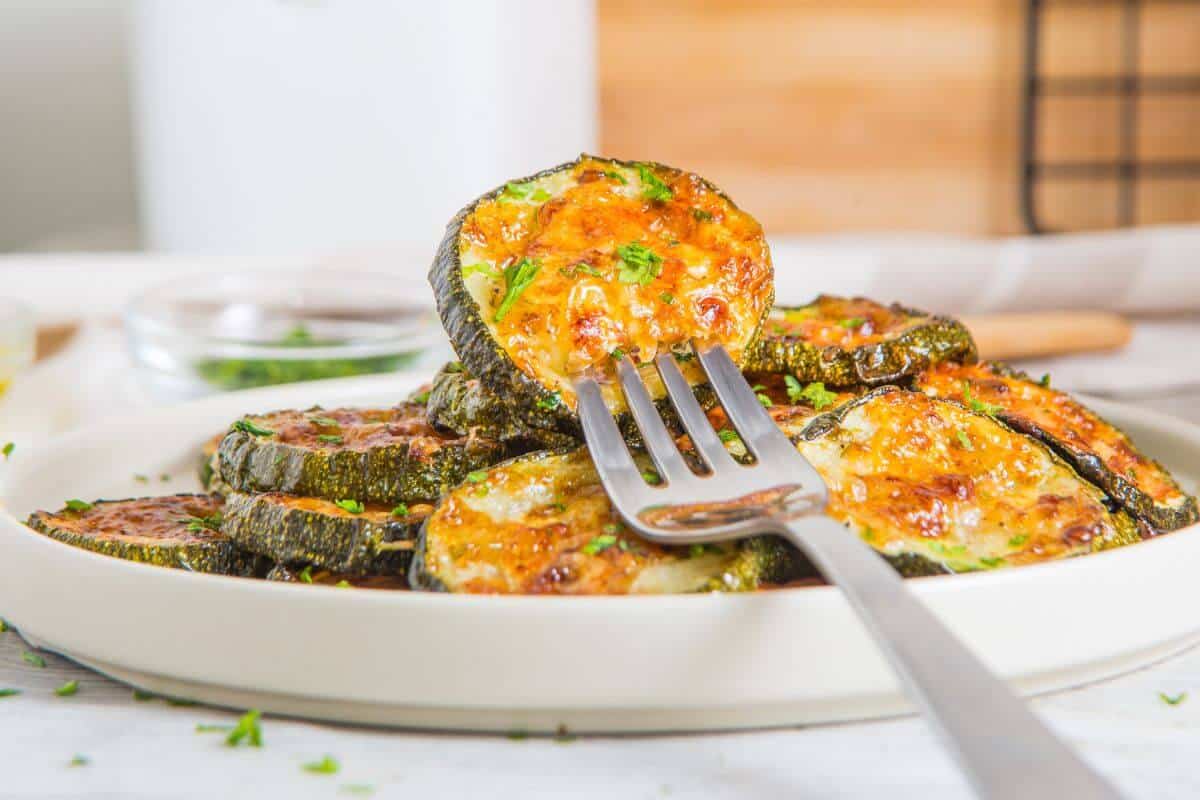 A close-up of a fork holding a slice of perfectly roasted zucchini with melted cheese and herbs, served on a pristine white plate with additional roasted zucchini slices elegantly arranged in the background.