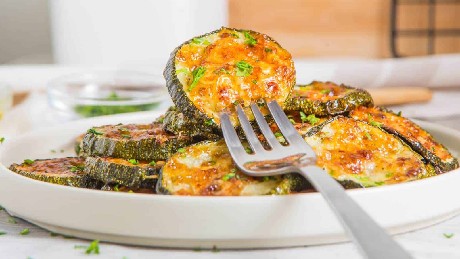 Roasted zucchini slices with melted cheese and herbs sit enticingly on a white plate, as a fork elegantly holds one piece.